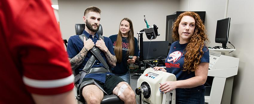 学生 working in exercise lab with one student in chair with testing equipment.